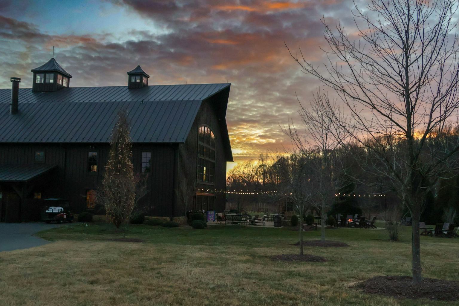Black board and batten barndominium with a standing seam metal roof and cupolas, windows glowing at sunset