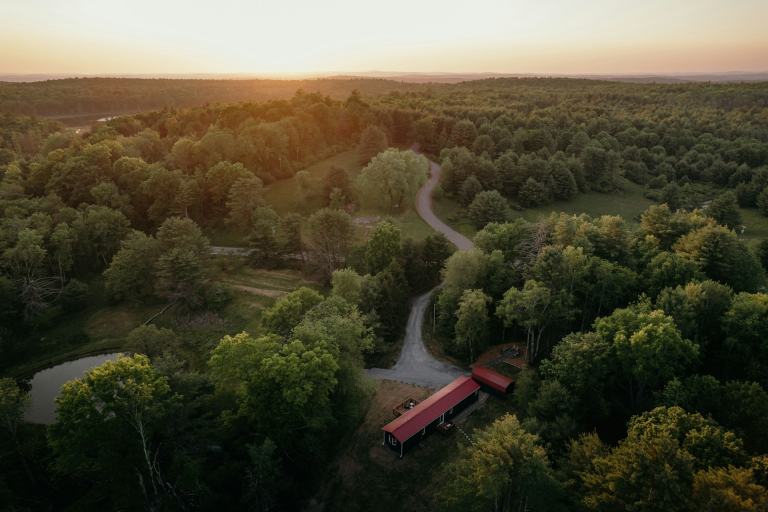 Aerial view of wooded rolling acreage with a pond, gravel drive and red-roofed outbuildings at golden hour, the kind of land found around Dickson, Tennessee