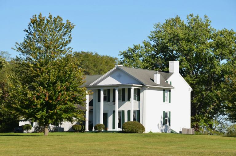 White two-story Southern farmhouse with columns and green shutters on a wide lawn under mature trees, the kind of home found around Lebanon and Wilson County, Tennessee