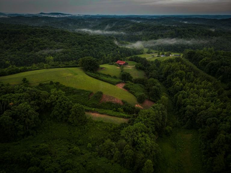Aerial view of wooded ridges and a hilltop pasture with exposed red clay soil under morning fog, the kind of terrain found across the Highland Rim around Nashville