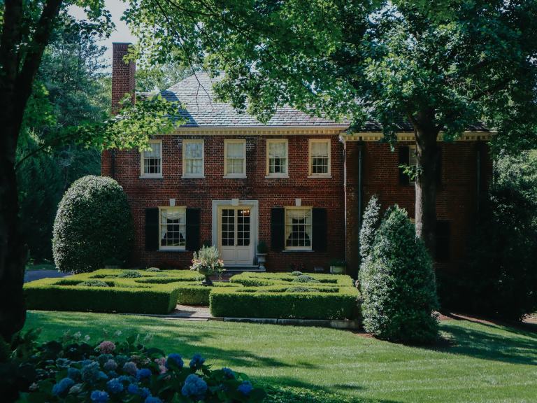 Red brick Georgian colonial home with black shutters, a boxwood parterre garden and blue hydrangeas under mature shade trees