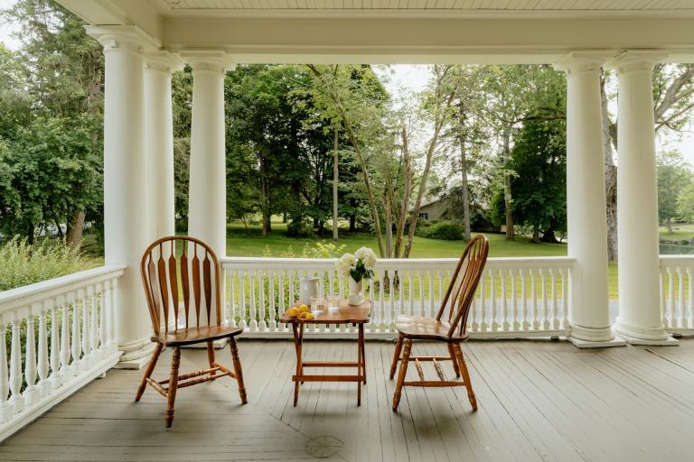 Wide covered front porch with white columns, two wooden chairs and a small table looking out over a green lawn and mature trees